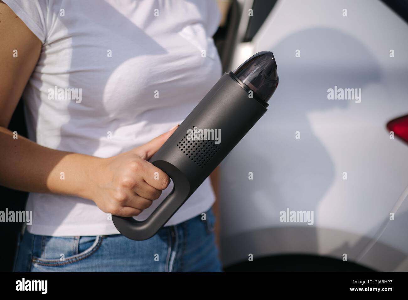 Femme tenir l'aspirateur portable devant la voiture. Belle jeune femme se prépare pour le nettoyage dans le garage. Gros plan des mains Banque D'Images