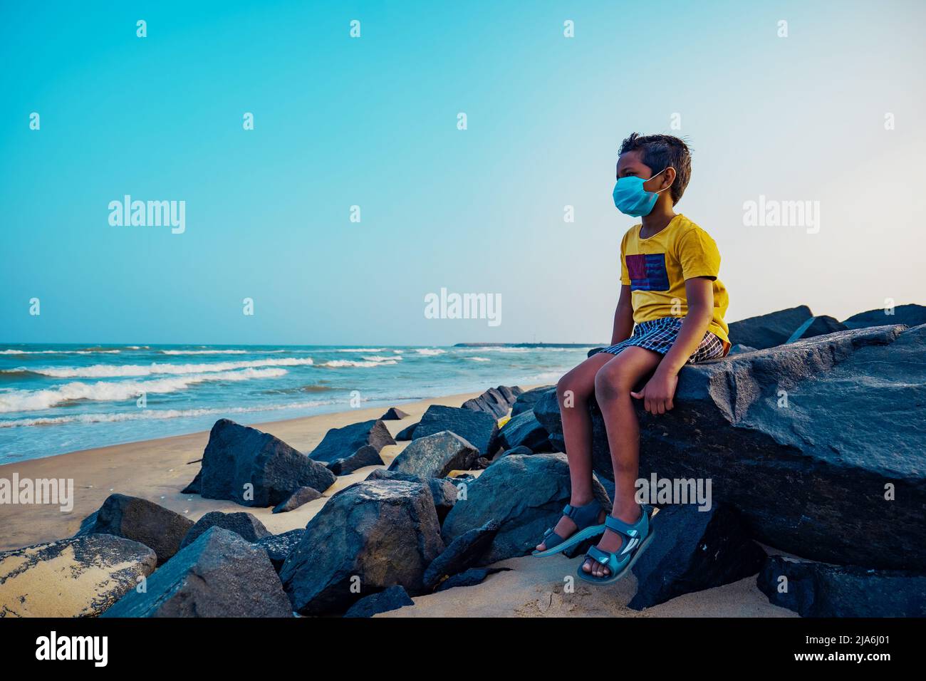 Garçon assis seul sur la plage avec un masque médical - garçon indien triste - garçon asiatique de 8-10 ans. Banque D'Images