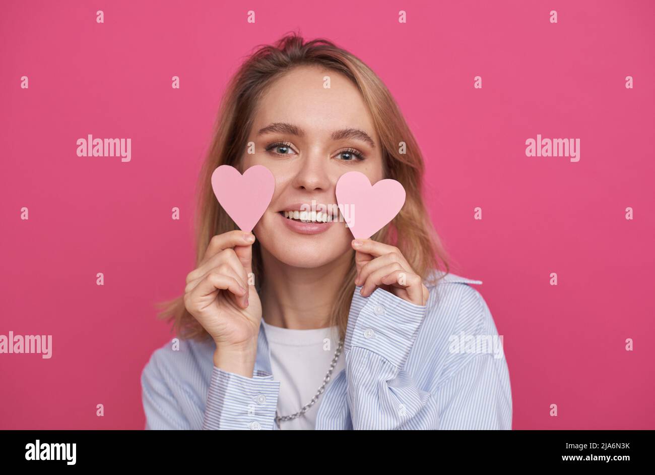 Portrait moyen en gros plan d'une belle jeune femme aux cheveux blonds debout contre un fond de mur rose tenant les coeurs en papier souriant à l'appareil photo Banque D'Images