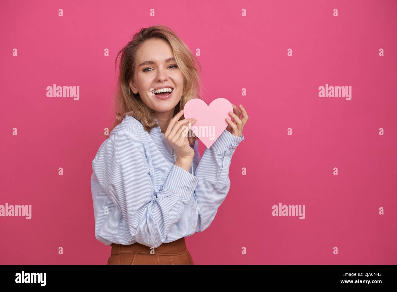 Portrait moyen d'une jeune femme joyeuse avec des cheveux blonds debout contre le fond de mur rose tenant le coeur de papier souriant à l'appareil photo Banque D'Images