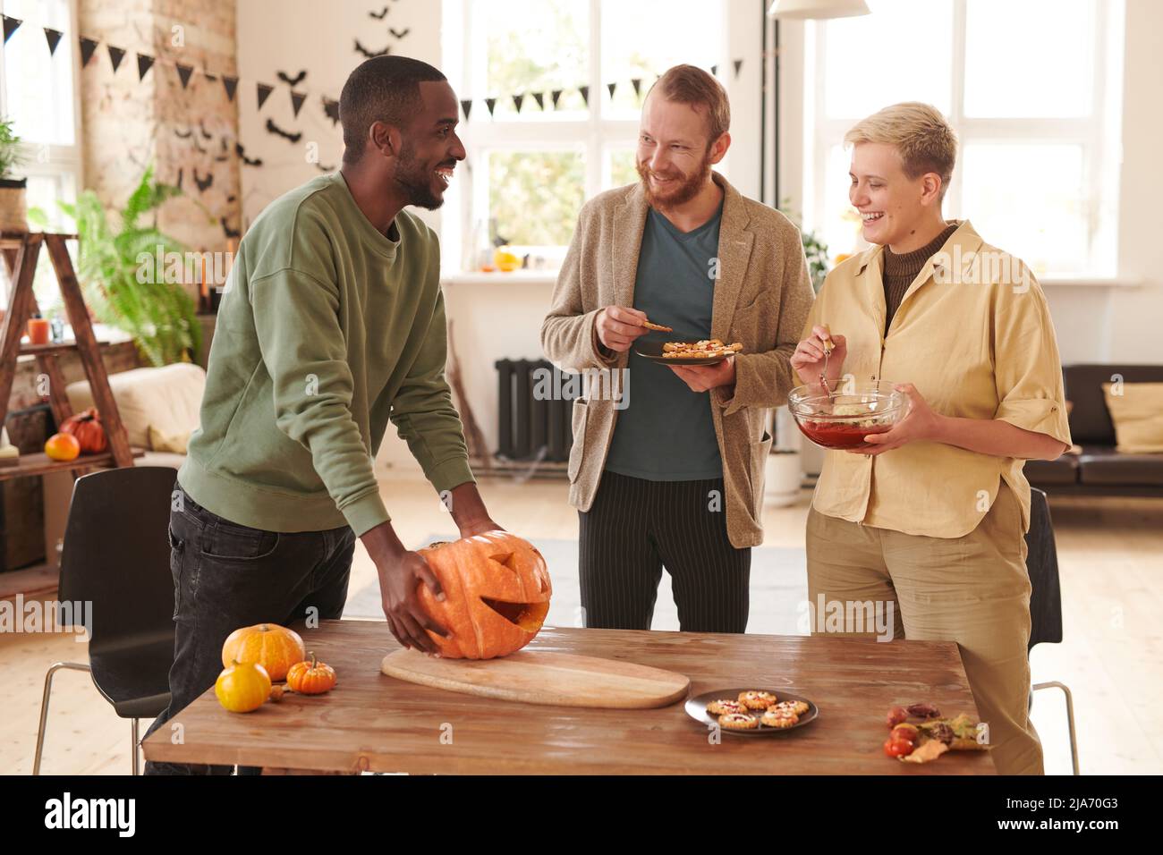 Groupe de jeunes amis multi-ethniques positifs dans des tenues décontractées debout sur une table en bois et faisant des gâteries pour Halloween partie Banque D'Images