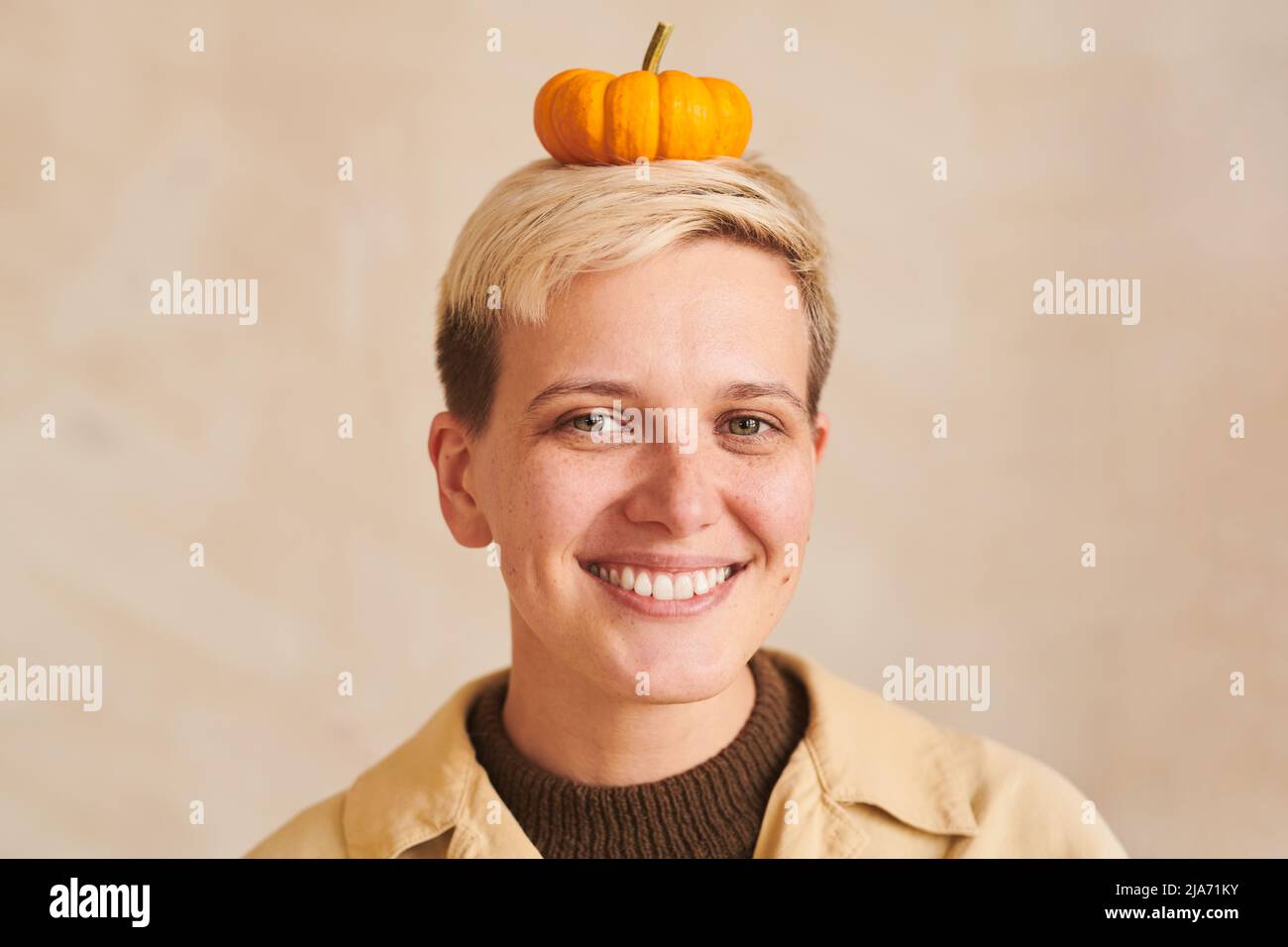 Portrait d'une jeune femme souriante et ludique avec des cheveux blonds tenant une petite citrouille mignonne sur la tête sur fond beige Banque D'Images