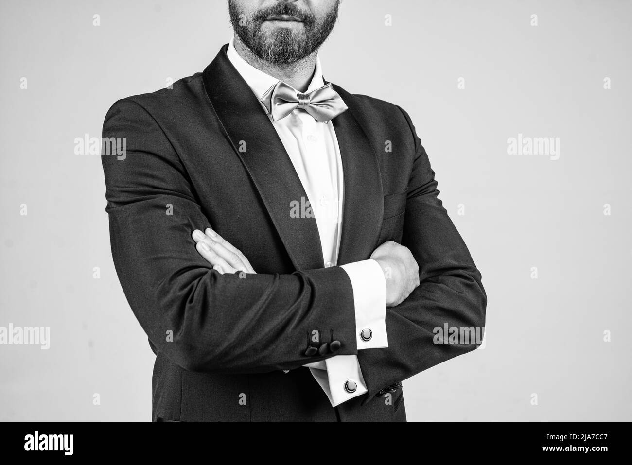 Visière confiante avec barbe non rasée coupe courte porter un smoking élégant avec noeud papillon en gardant les bras croisés fond gris, confiance Banque D'Images