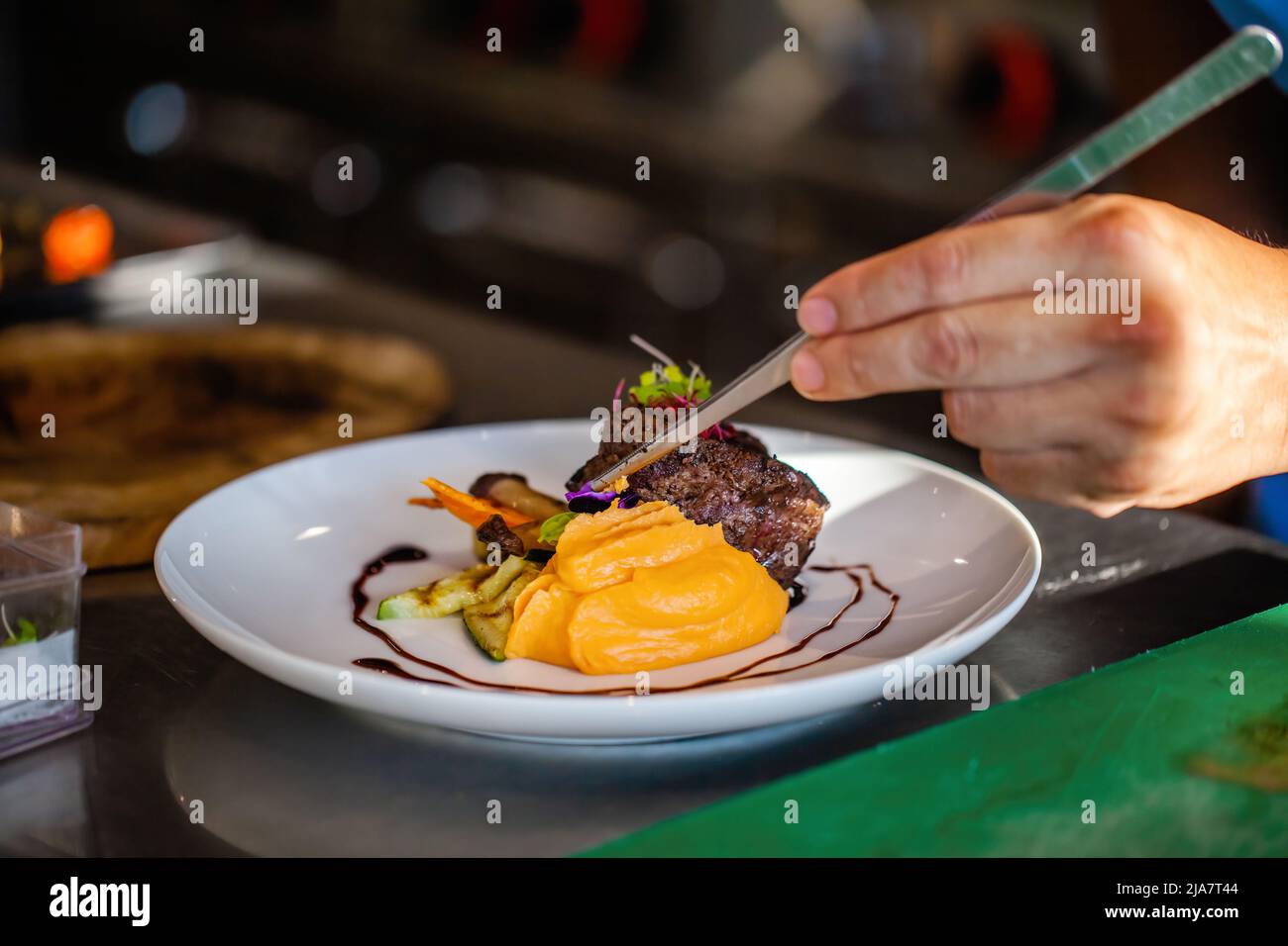 Gros plan des mains du chef dans la cuisine de l'hôtel ou du restaurant décorant le plat. Préparer le steak de bœuf avec la purée de pommes de terre, ajouter les légumes, décorer le plat avec Banque D'Images