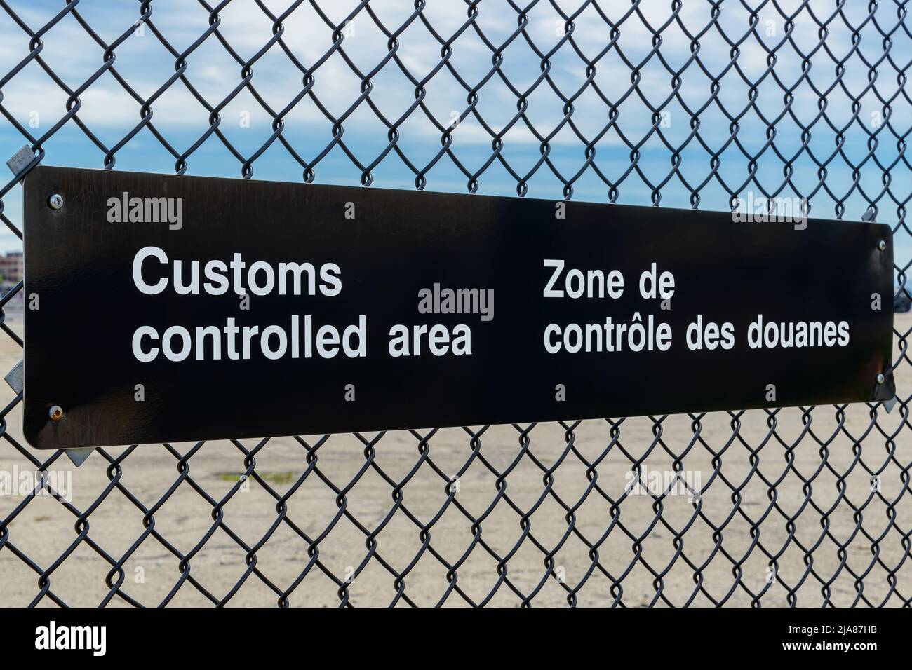 Une affiche bilingue de zone contrôlée par les douanes en anglais et en français sur une clôture à chaînette. Vue en angle avec ciel bleu au-dessus, parking en dessous. Banque D'Images