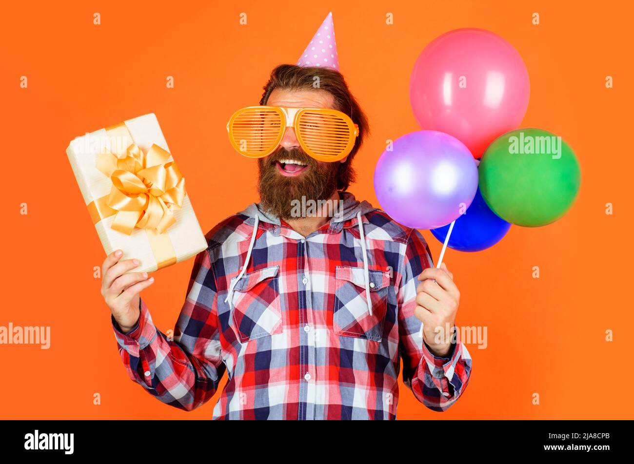 Joyeux anniversaire. Homme à barbe souriant dans un chapeau de fête et des lunettes avec des ballons et un cadeau. Banque D'Images