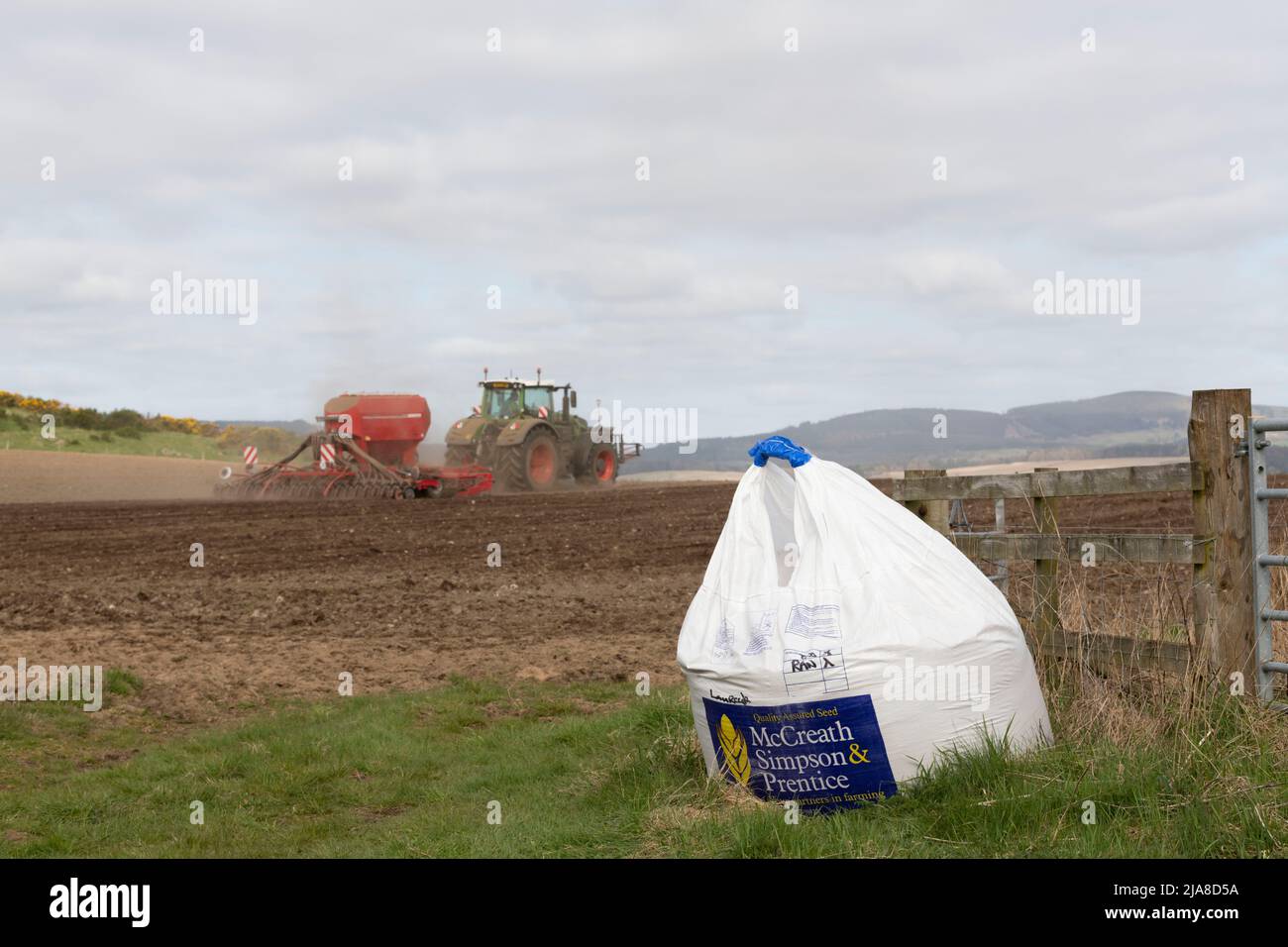 Un sac de semences du commerçant assis à côté de l'entrée d'un champ labouré avec un tracteur vert tractant un semoir fonctionnant en arrière-plan Banque D'Images