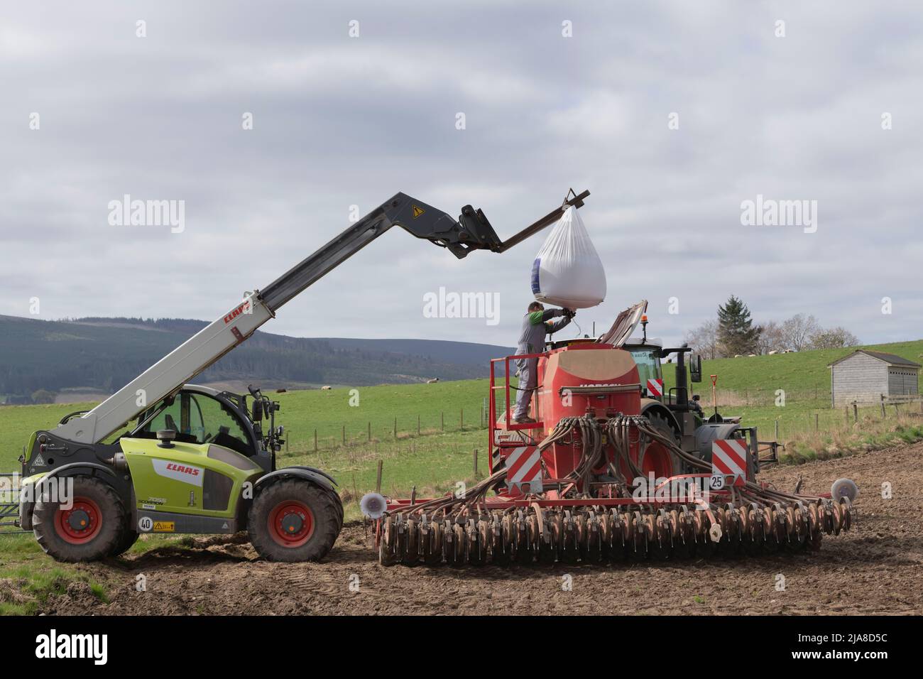 Un agriculteur qui remplit un semoir universel Horsch à partir d'un sac de semences du marchand suspendu d'un chargeur à bras télescopique Claas dans un champ labouré Banque D'Images