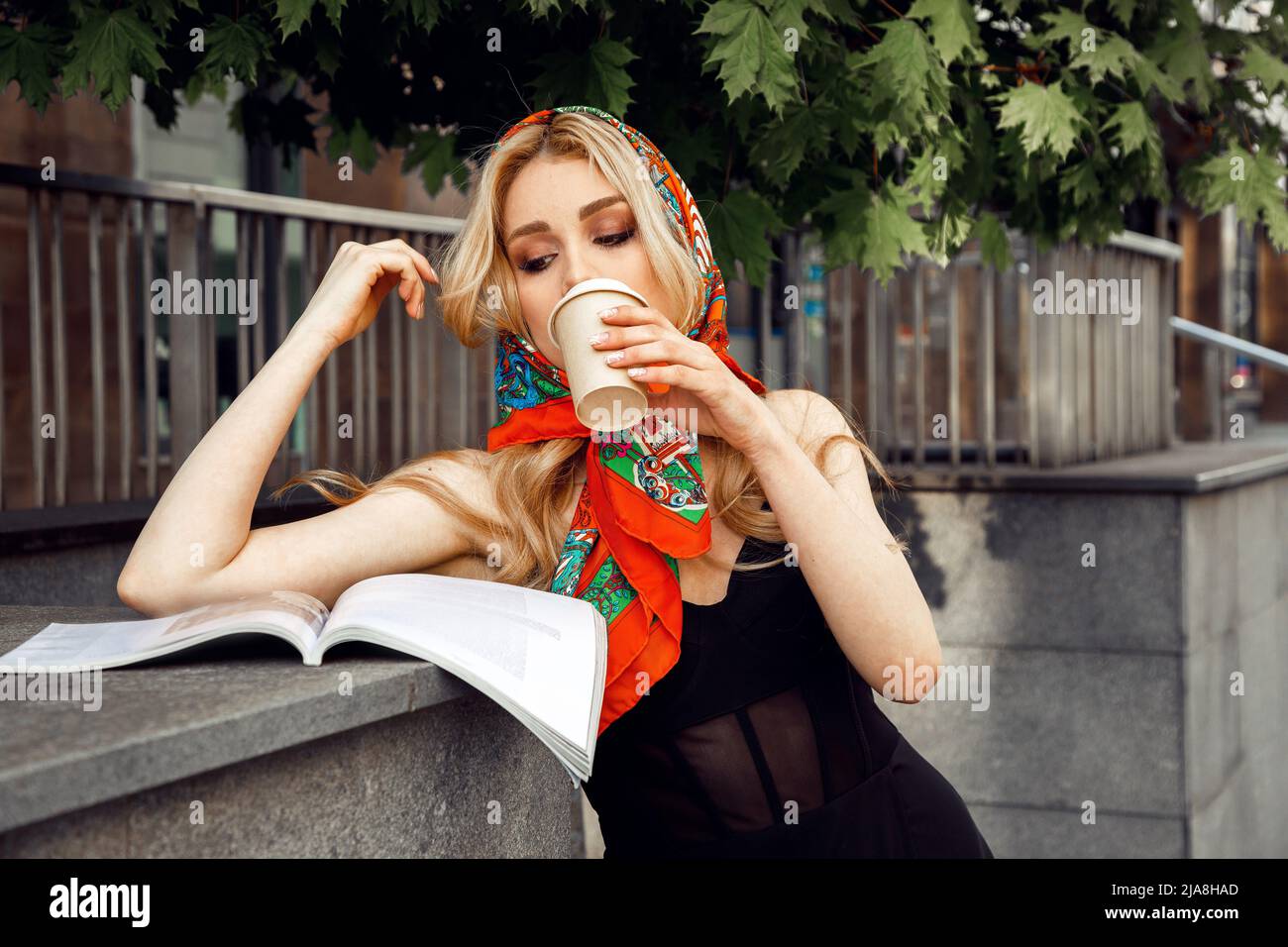 Jeune femme portant une robe noire, un mouchoir en soie, debout sur la main courante, feuilletant le magazine, buvant une tasse de café. Banque D'Images