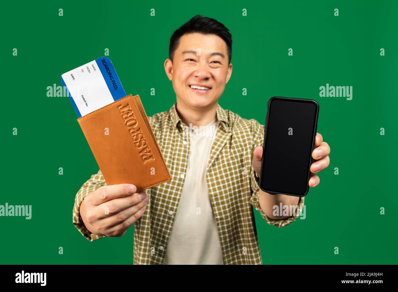 Joyeux homme asiatique tenant un téléphone portable avec écran vierge et un passeport avec des billets, posant sur fond vert Banque D'Images