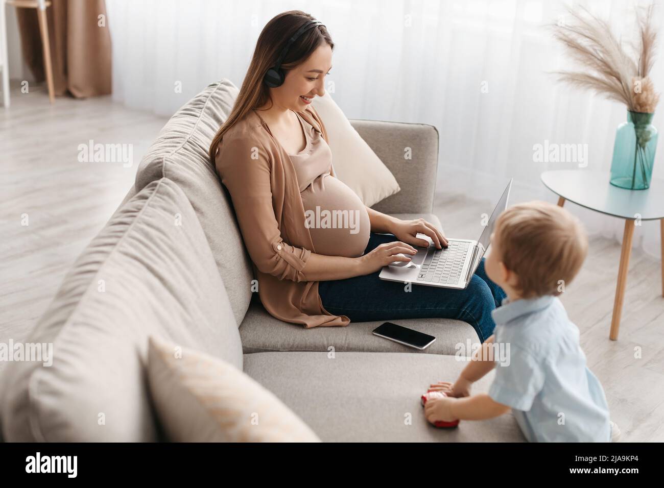 Bonne femme enceinte dans un micro-casque utilisant un ordinateur portable, étudiant ou travaillant en ligne pendant que le petit fils joue à proximité Banque D'Images