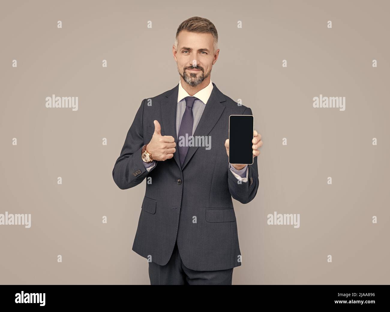 homme mature influenceur en costume présentant l'appareil de téléphone mobile et montrer le pouce vers le haut, meilleure offre. Banque D'Images