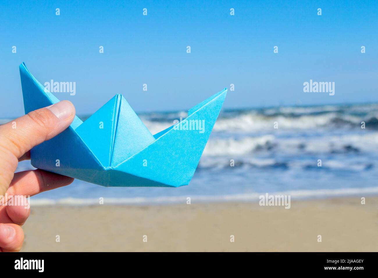 Personne tenant le bateau en papier bleu à la main et jouant flottant dans l'air sur fond vagues de mer. Ciel bleu et plage de sable sur le soleil jour d'été concept tourisme vacances repos rêve voyage voile Banque D'Images