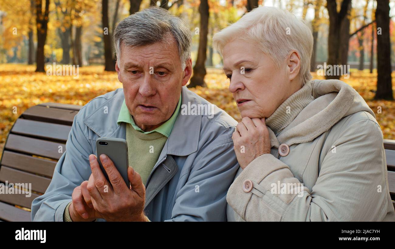 Contrarié vieux marié couple caucasien utiliser smartphone s'asseoir sur le banc en automne parc regarder écran téléphone inquiet personnes âgées retraités triste sentiment de famille Banque D'Images