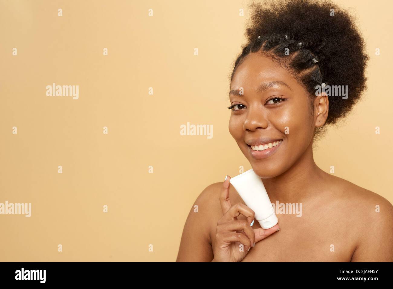 Belle femme afro-américaine tenant tube avec hydratant ou masque maquette sur fond beige avec espace de copie. Soins de la peau, anti-âge. Banque D'Images