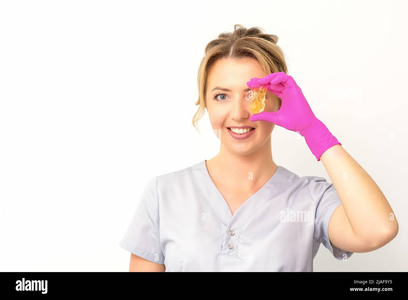 Le maître de l'enlèvement de cheveux de sucre tient la pâte de sucre jaune liquide, couvrant son oeil avec de la cire pour l'épilation sur un fond blanc Banque D'Images