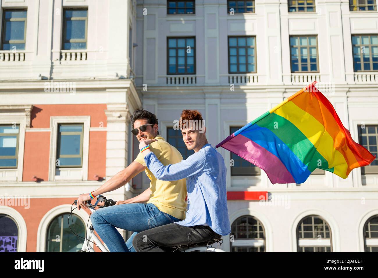 Jeunes hommes gay couple à vélo tenant gay fierté drapeau dans la ville regardant la caméra. concept lgbt Banque D'Images
