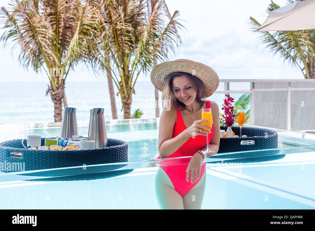 Femme de voyage souriante ou influenceur buvant du jus d'orange dans la piscine transparente à débordement de l'hôtel de luxe. Vacances d'été exotiques sur la plage Banque D'Images