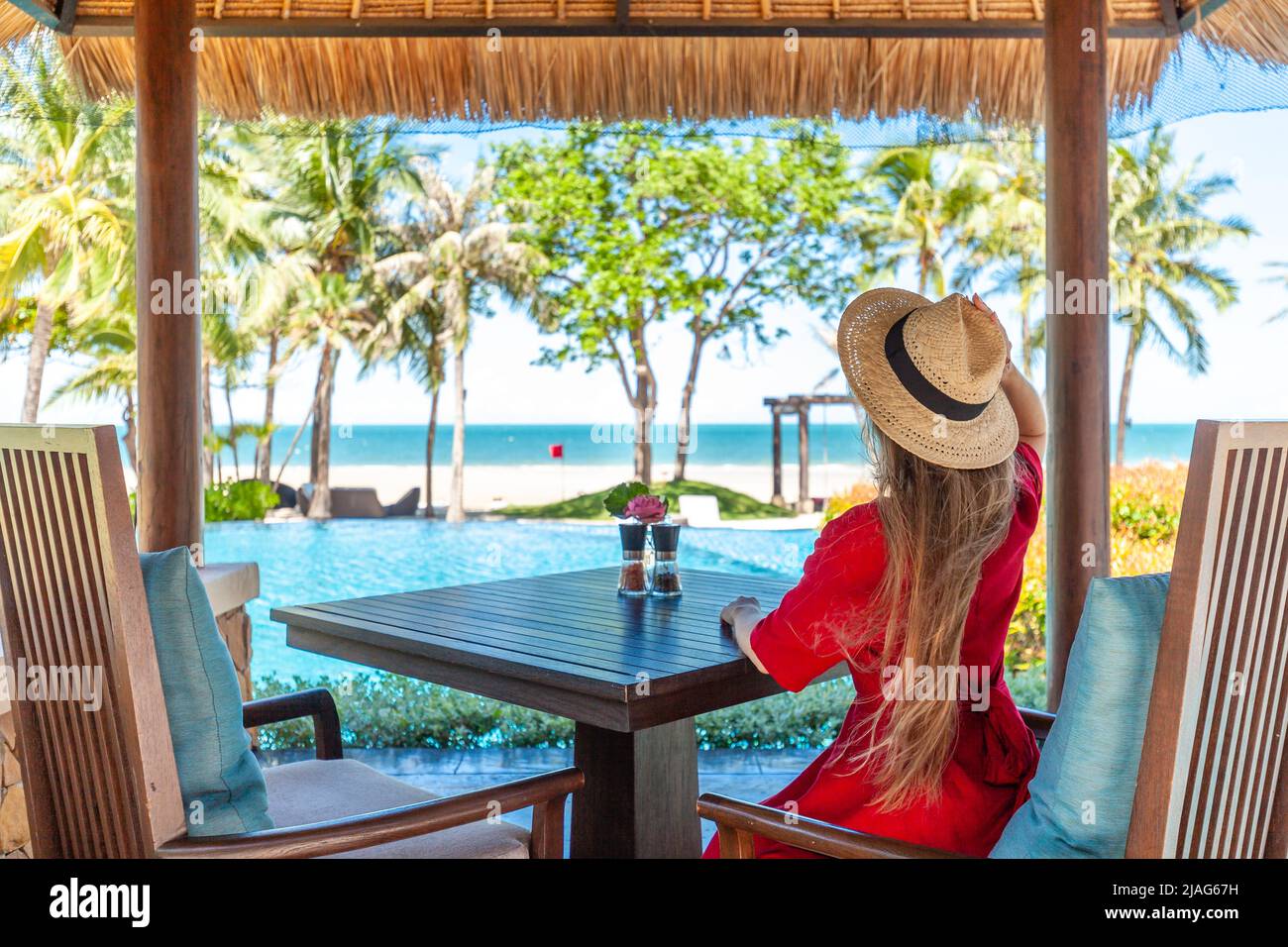 Femme en vacances d'été. Une fille de voyage vêque de rouge et chapeau assis sur la table du salon de l'hôtel près de la piscine. Restaurant de plage tropical avec Banque D'Images