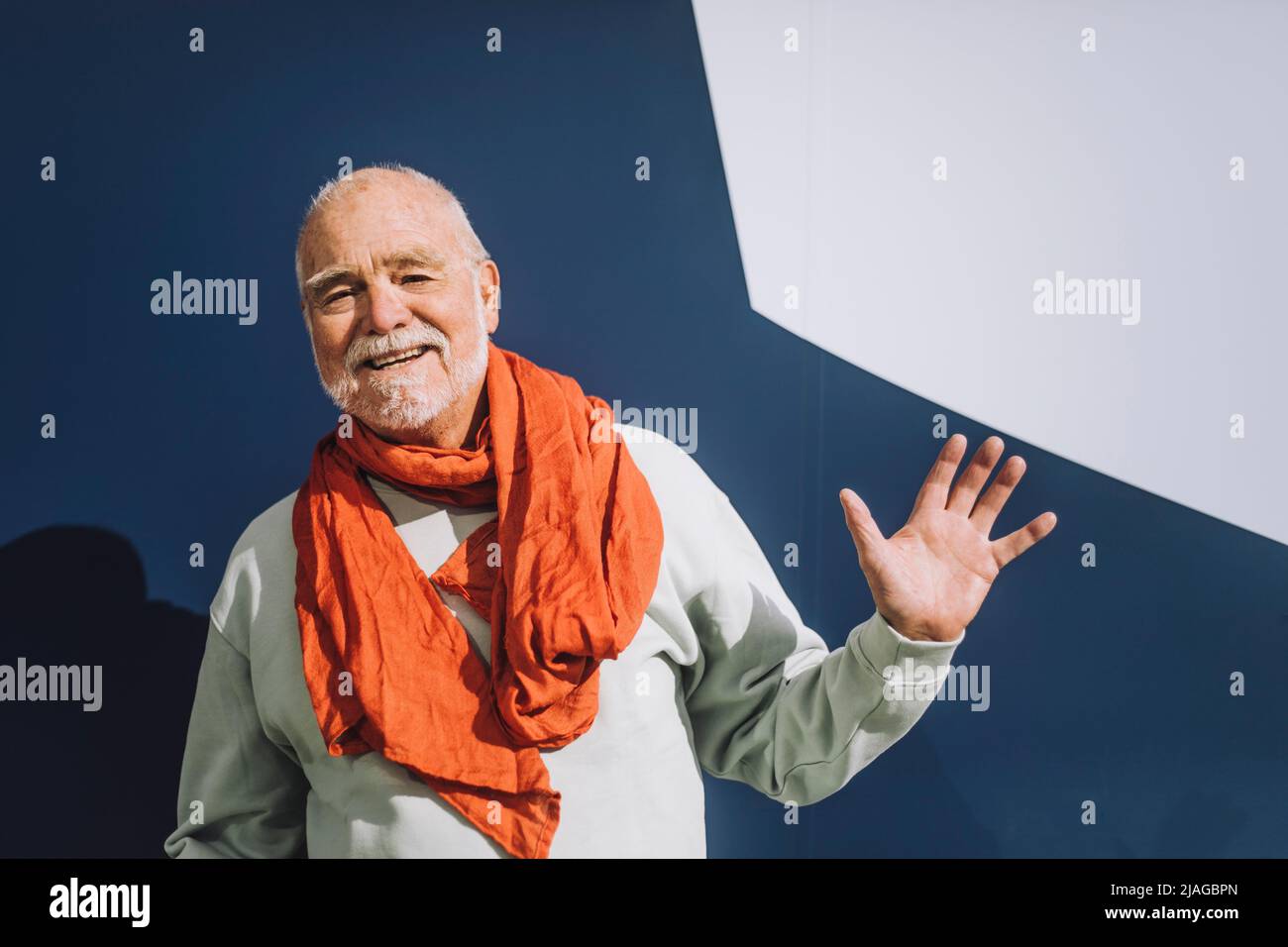 Portrait d'un homme âgé heureux portant un foulard qui agite la main contre un mur bleu et blanc Banque D'Images