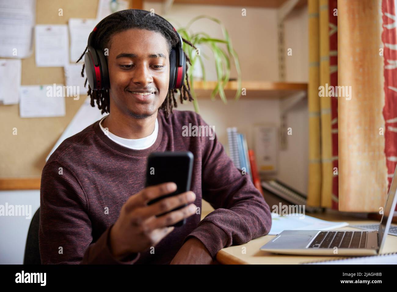 Un étudiant d'université ou d'université travaillant sur un ordinateur portable portant un casque sans fil regardant un téléphone portable au bureau dans la chambre Banque D'Images