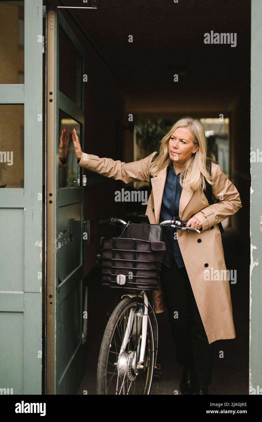 Femme d'affaires portant un long manteau ouvrant la porte tout en tenant le vélo Banque D'Images