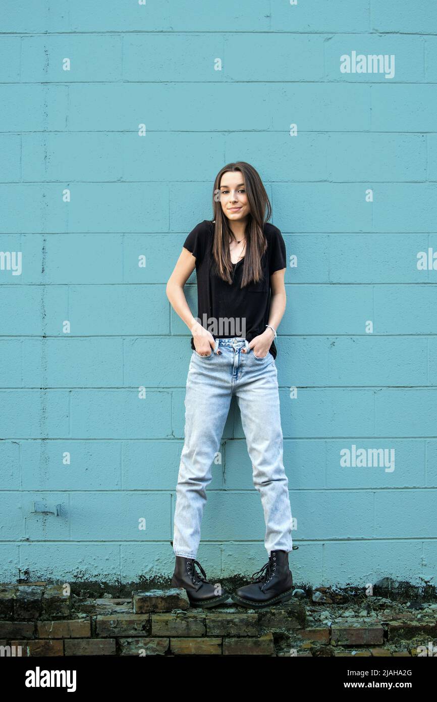 Une adolescente aux cheveux longs portant des chaussures noires, en denim et de combat, penchée contre un mur de blocs de cinder bleu et regardant loin avec l'attitude Banque D'Images
