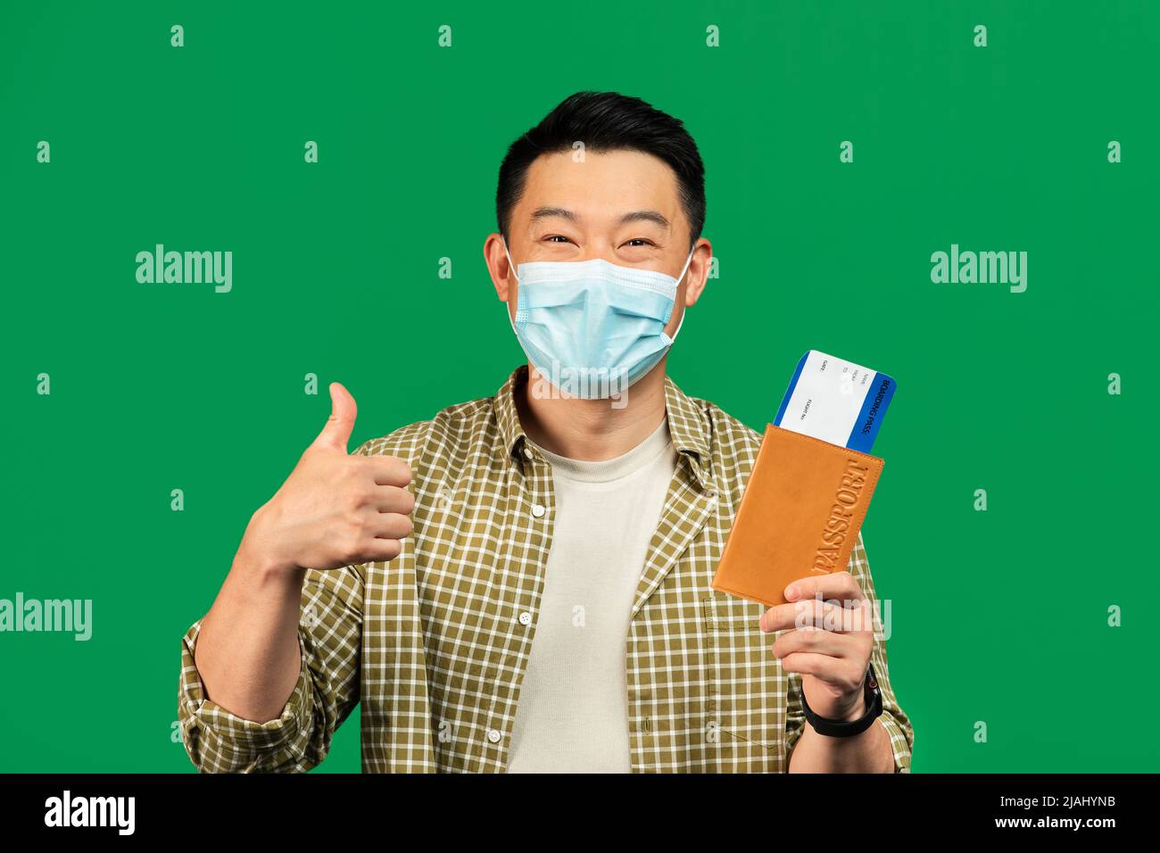 Portrait d'un touriste asiatique mature, masque, passeport avec billets, pouce vers le haut, fond vert Banque D'Images