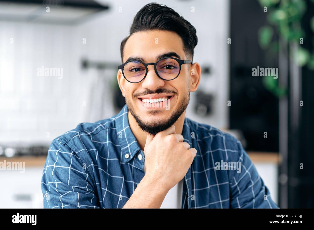 Gros plan d'un jeune garçon arabe ou indien positif avec des lunettes, un indépendant ou un étudiant, dans des vêtements décontractés, avec des dents blanches parfaites, regardant l'appareil photo avec un sourire joyeux et ensanglant Banque D'Images