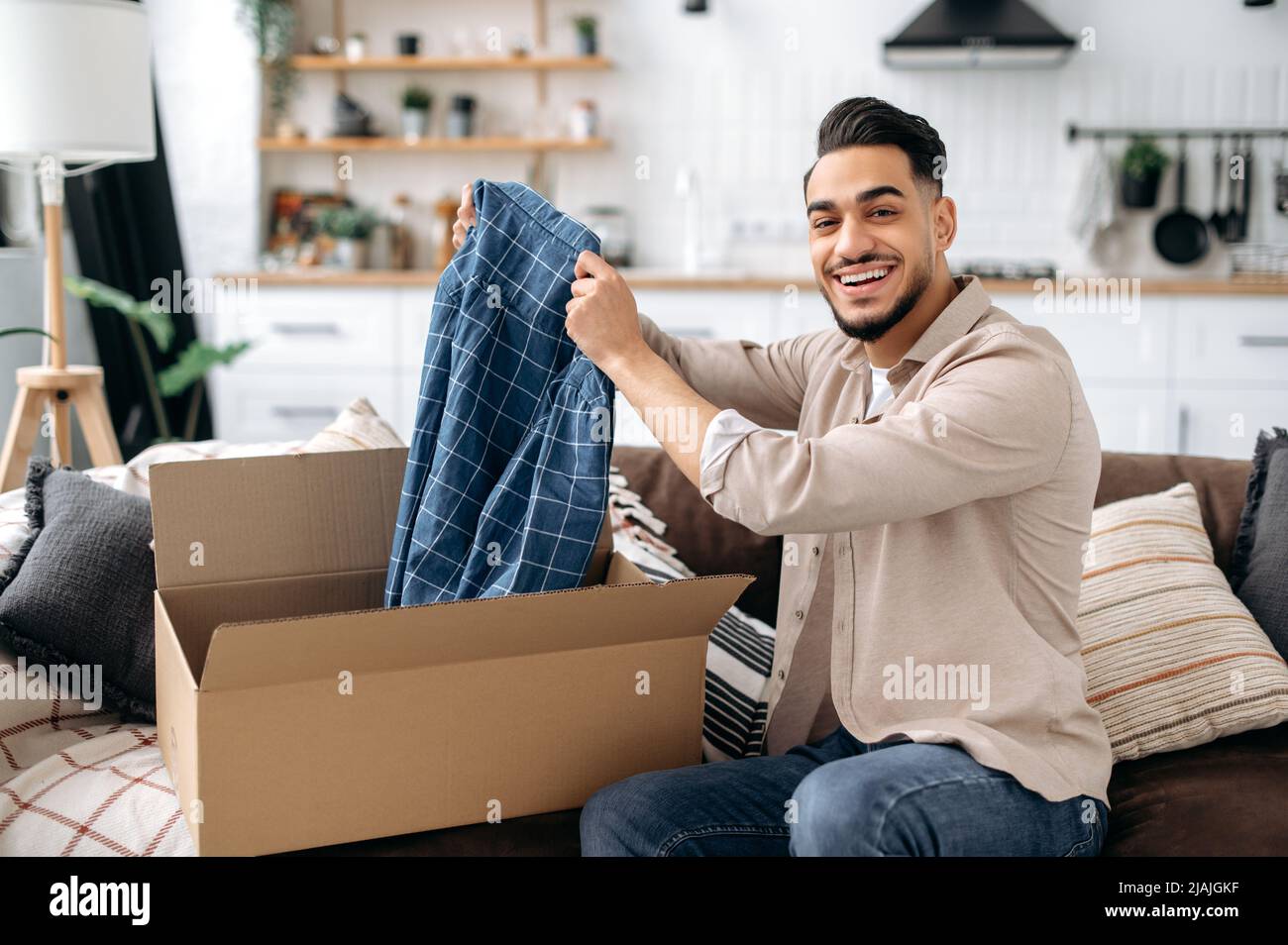 Beau positif indien ou arabe gars ouvrant grande boîte de carton, déballage de la commande de magasin d'Internet à la maison, se sentent satisfaits avec le service de livraison rapide et les marchandises qui ont reçu, sourire heureux Banque D'Images