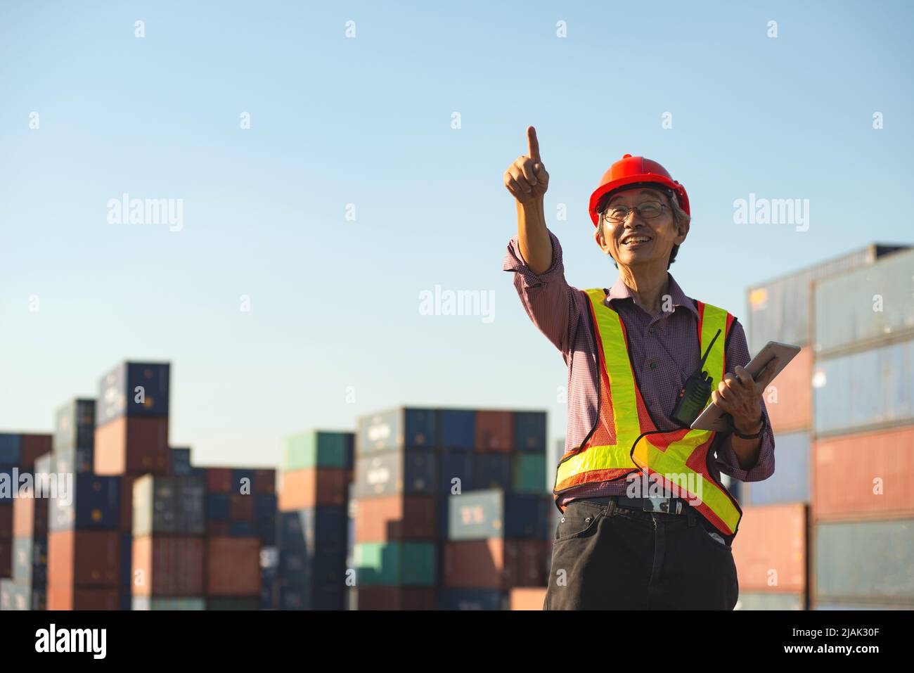 Un ingénieur travailleur asiatique âgé de longue date portant un gilet de sécurité et un casque debout et tenant une tablette numérique dans la cour d'expédition des conteneurs de fret. Banque D'Images