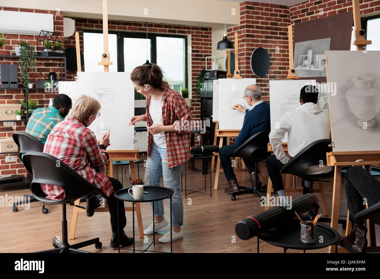 Jeune enseignante aidant la femme âgée à développer la technique d'illustration tout en assistant à la leçon artistique pour la croissance personnelle. Groupe multiethnique d'étudiants dessinant un modèle de vase sur toile blanche Banque D'Images