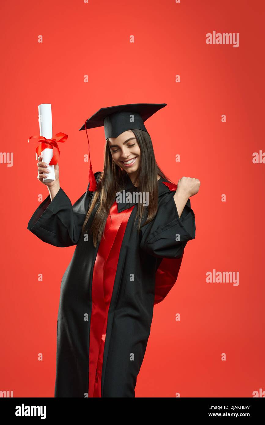 Vue de face d'une jeune fille heureuse avec baccalauréat, diplôme de maîtrise. Jolie femme debout avec les yeux fermés, montrant le hurray, souriant. Isolé sur fond rouge studio. Banque D'Images Vue de face d'une jeune fille heureuse avec baccalauréat, diplôme de maîtrise. Jolie femme debout avec les yeux fermés, montrant le hurray, souriant. Isolé sur fond rouge studio. Banque D'Images