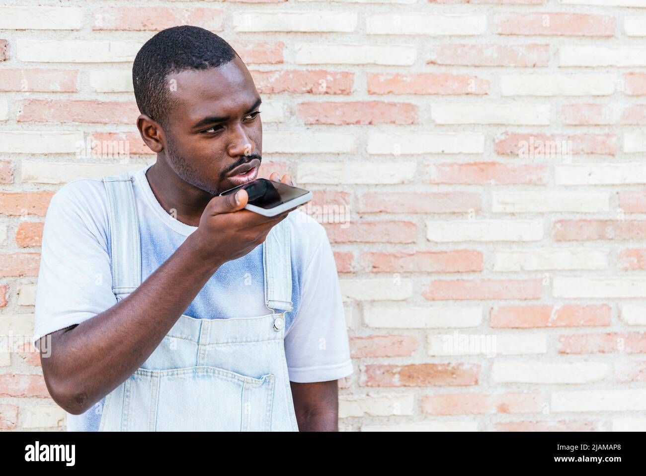 Un jeune homme afro-américain sérieux portant des vêtements décontractés. Il enregistre des messages vocaux sur son smartphone et regarde loin tout en se tenant près du mur de briques sur la rue de la ville Banque D'Images