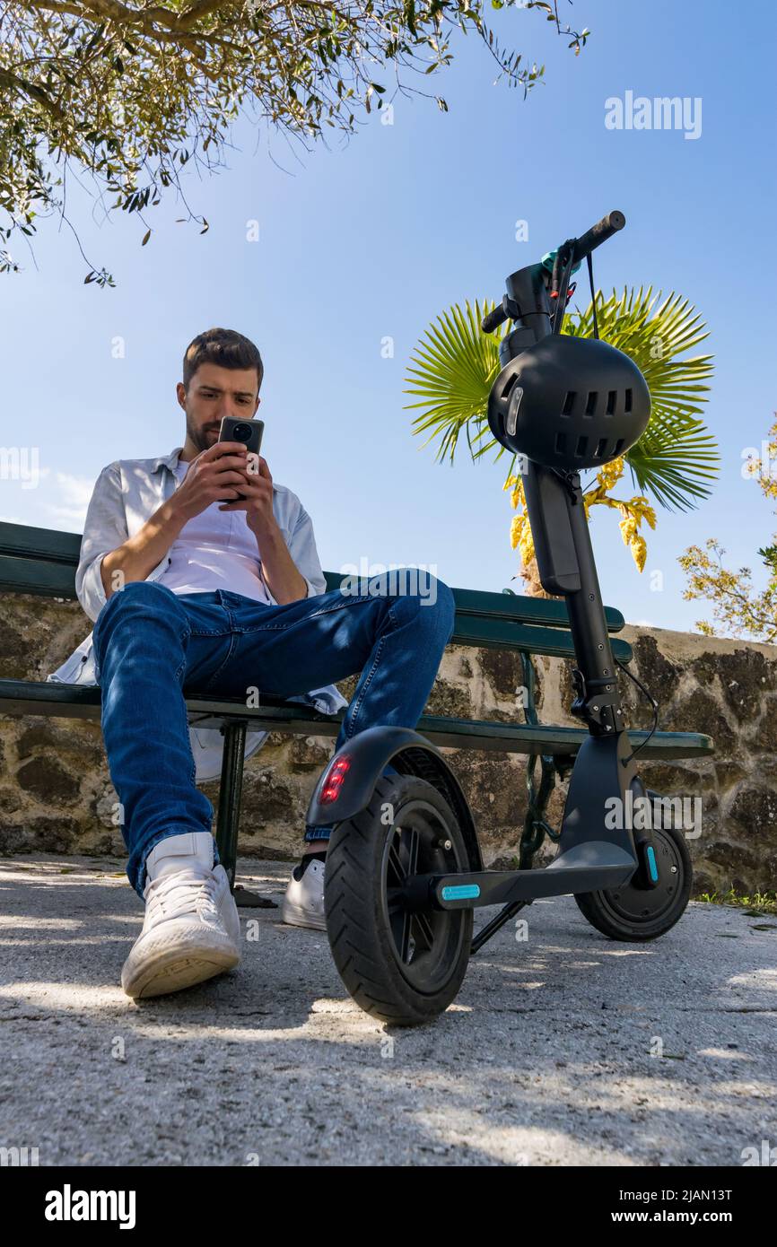 Le jeune homme regarde son smartphone assis sur un banc de parc à côté de son scooter électrique Banque D'Images
