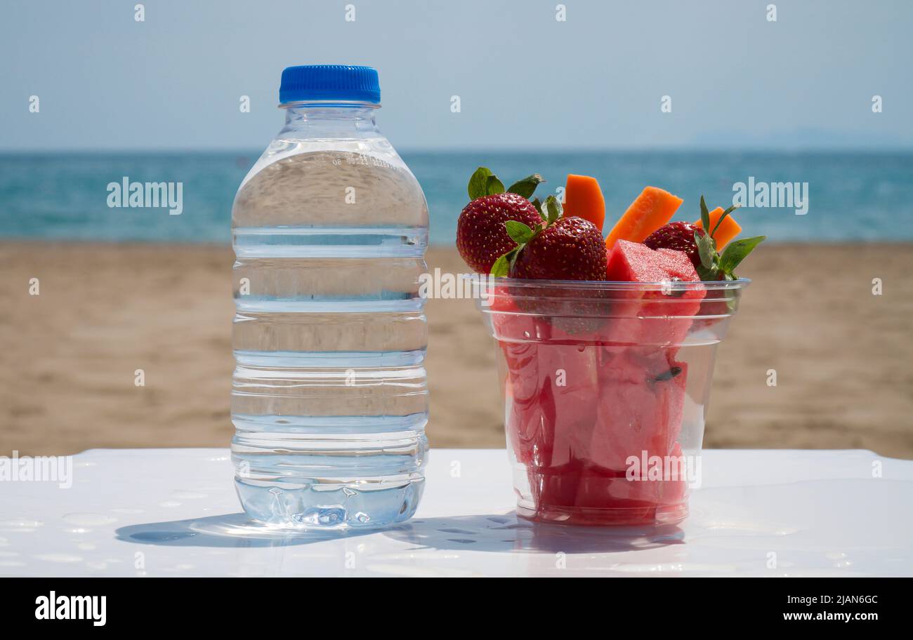 Bouteille d'eau en plastique et fruits sur une tasse en plastique, sur une table blanche. Toile de fond de la plage Banque D'Images
