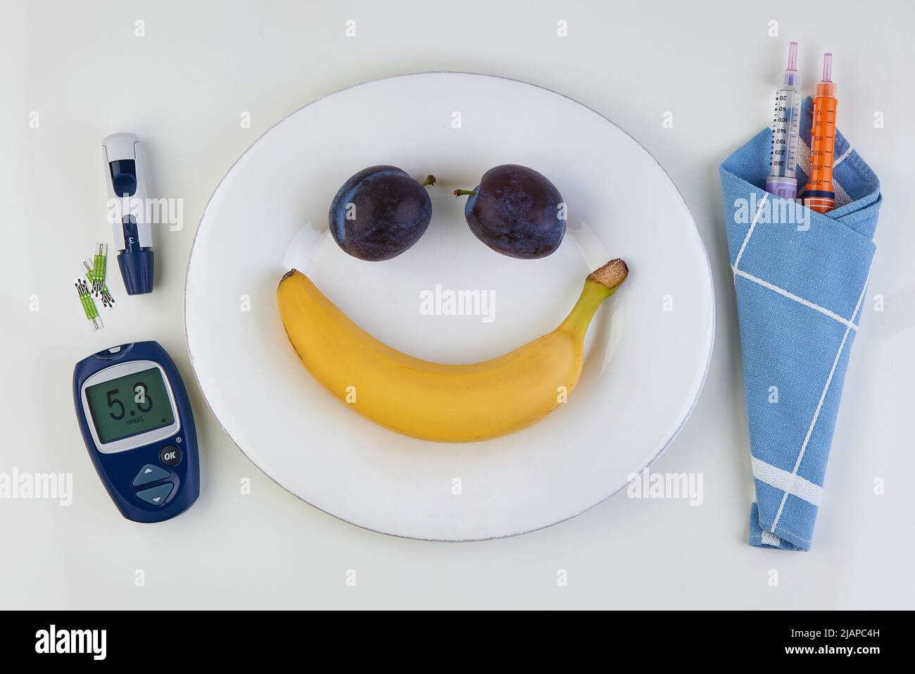 Banane et prune sous la forme d'une émoticône souriante sur une plaque blanche et un stylo pour seringues d'insuline et un glucomètre comme coutellerie sur une table blanche. Banque D'Images