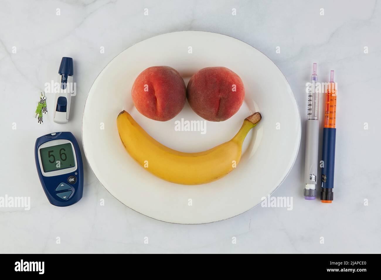 Banane et pêches sous la forme d'une émoticône souriante sur une plaque blanche et des stylos à seringue d'insuline et un glucomètre comme couverts sur une table en marbre. Banque D'Images