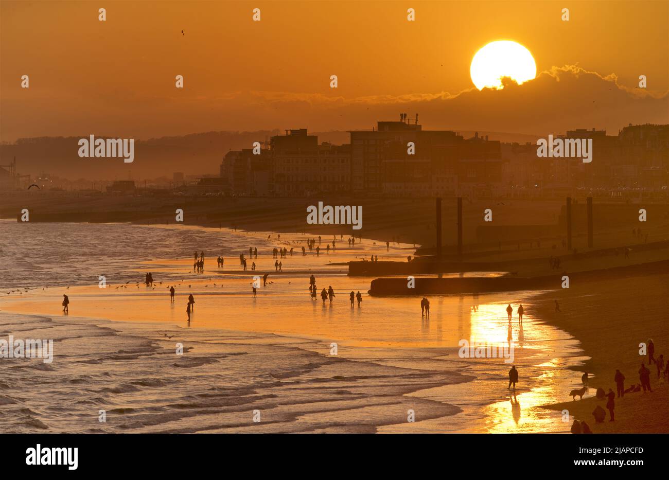 Silhoueted formes de personnes sur la plage à marée basse, Brighton & Hove, East Sussex, Angleterre, Royaume-Uni. Le soleil se couche derrière le nuage. Piliers verticaux en fonte de l'ancien West Pier sur la droite. Banque D'Images