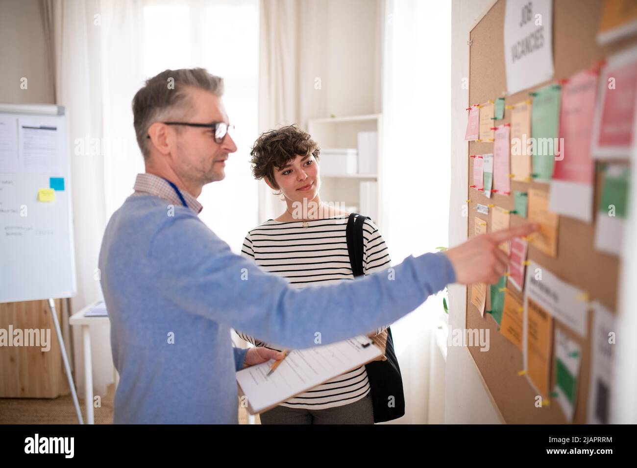 Employé de l'agence de recrutement debout devant le tableau d'affichage de l'emploi et aidant la jeune femme à rechercher un emploi. Banque D'Images