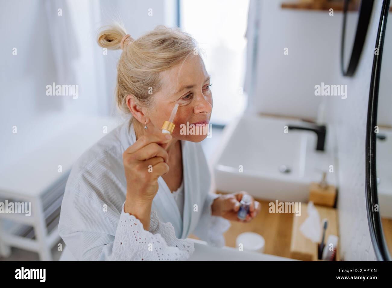 Belle femme sénior en peignoir, hydratant la peau par pipette avec du sérum appréciant la procédure de traitement. Banque D'Images