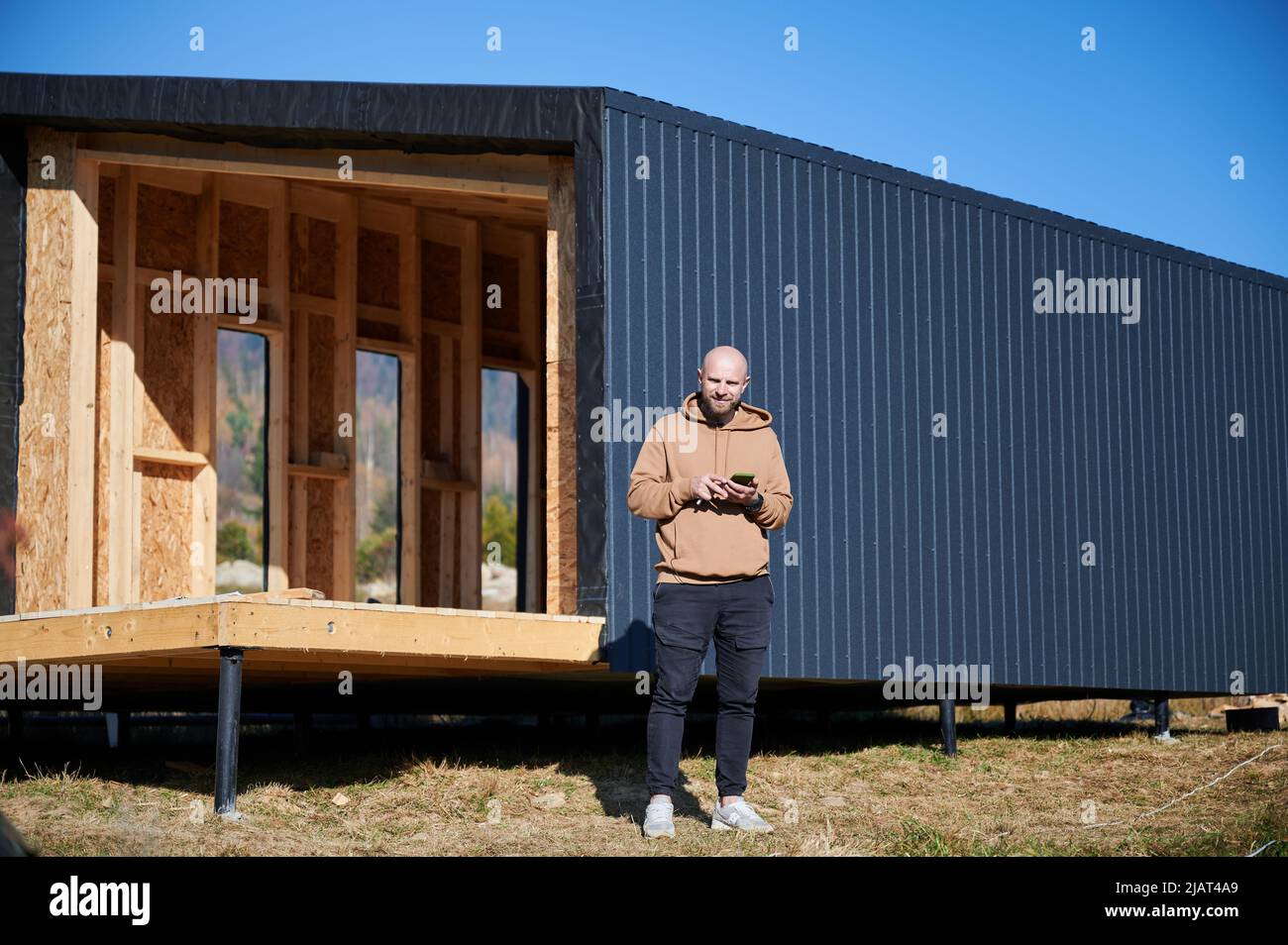 Maison d'architecte mâle en bois sur une fondation en pieu dans la maison de style scandinave. Portrait d'homme chauve debout sur le chantier avec téléphone, inspectant la qualité du travail. Banque D'Images