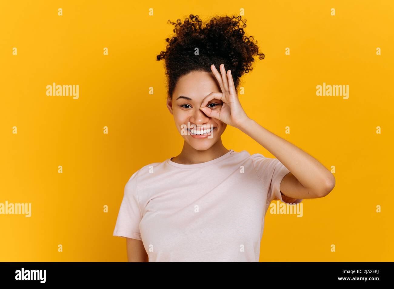 Bonne charmante jeune femme afro-américaine aux cheveux maurires, fait avec le doigt le symbole OK près de son œil, montre le signe d'accord, debout sur un arrière-plan orange isolé, regarde la caméra, souriant Banque D'Images