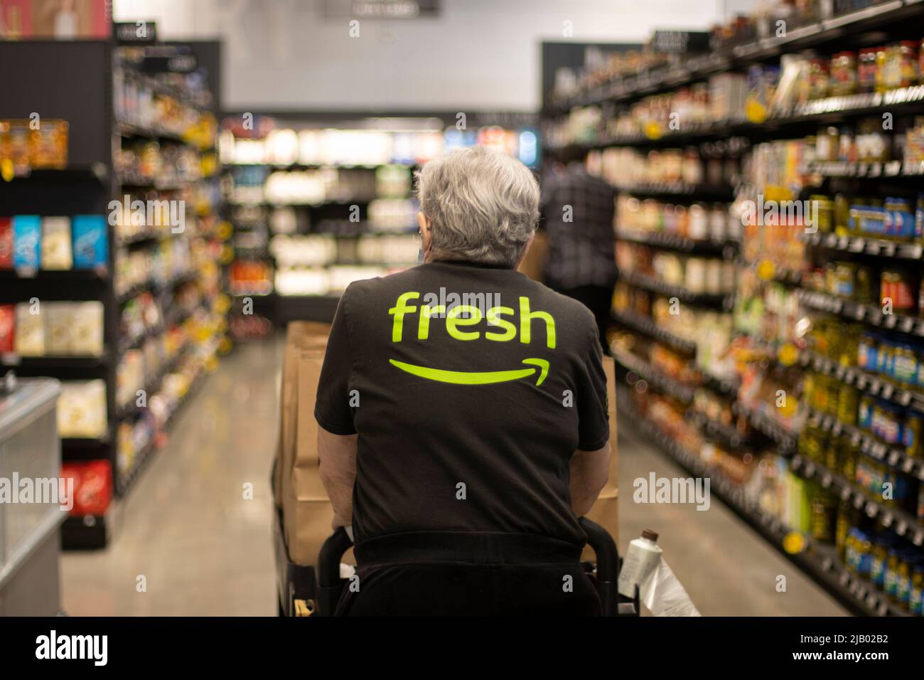 Un travailleur âgé dans un T-shirt frais Amazon pousse un chariot chargé de sacs de papier de ramassage dans les allées à l'intérieur d'un magasin d'épicerie frais Amazon en ... Banque D'Images