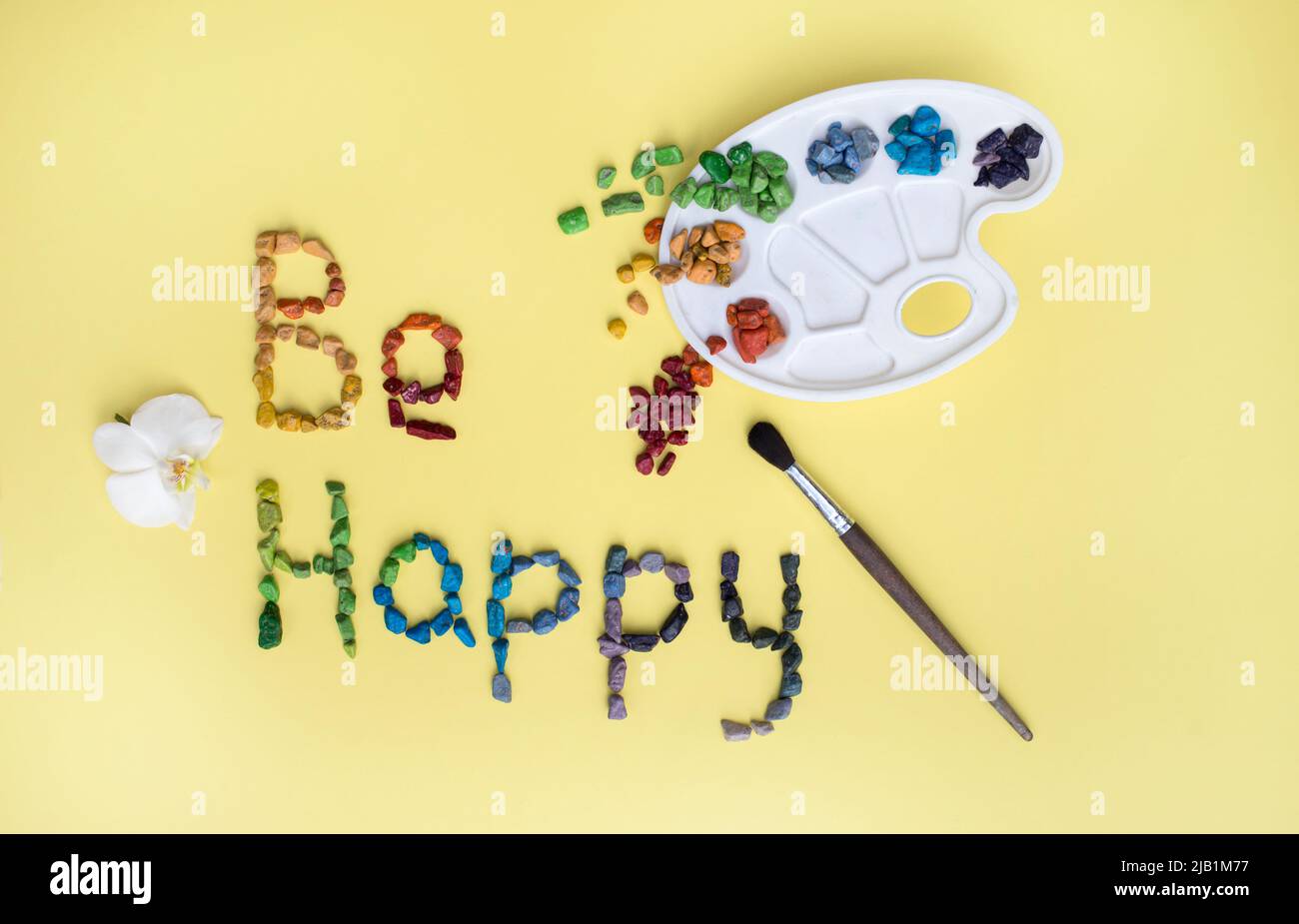 le désir d'être heureux est écrit à partir de bonbons multicolores sous forme de galets de mer, une palette pour aquarelle, un pinceau sur fond jaune. Anniversaire Banque D'Images