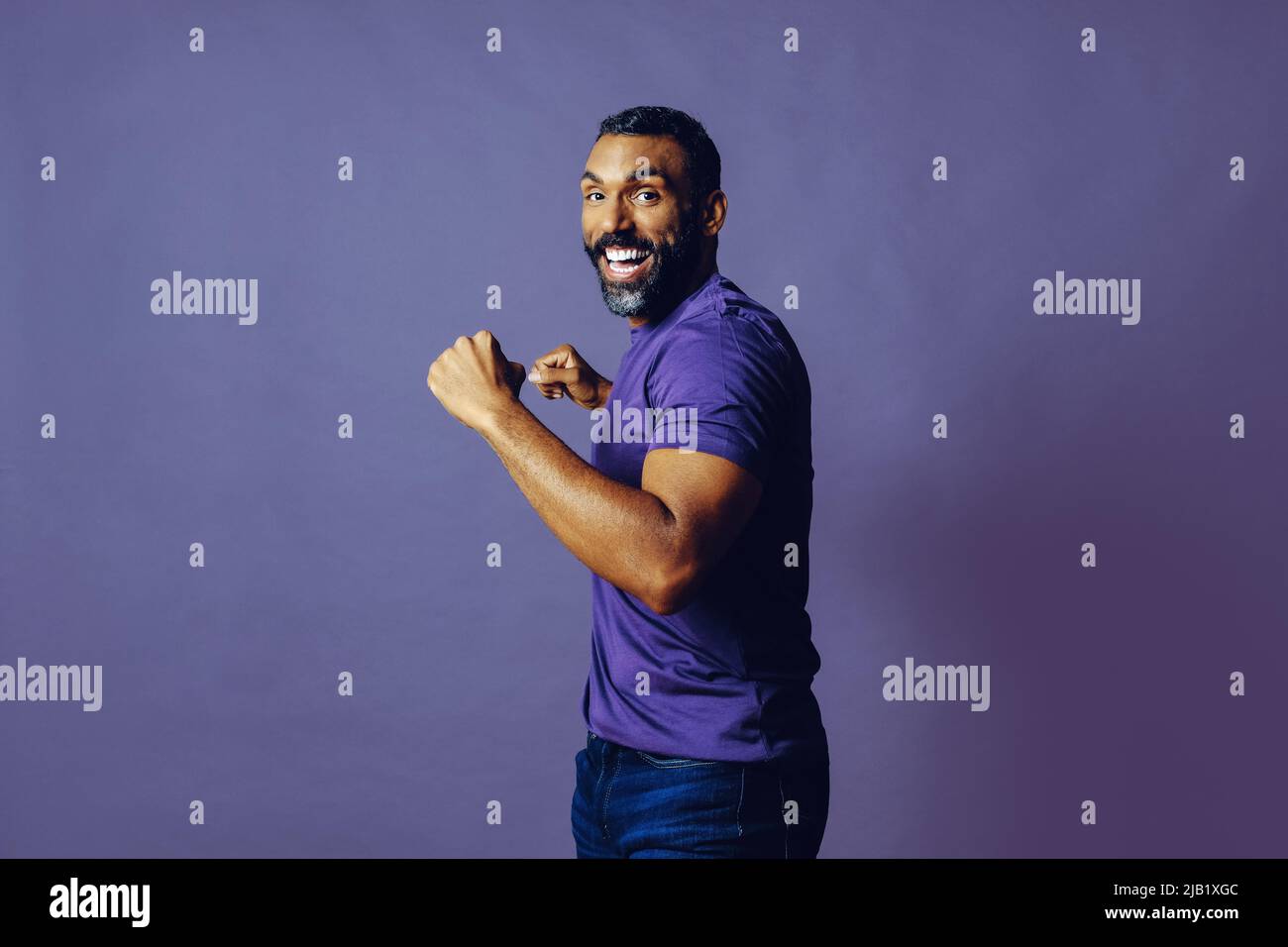 portrait d'un homme gagnant réussi avec une barbe et un t-shirt violet célébrant avec bras vers le haut sur un fond violet studio Banque D'Images