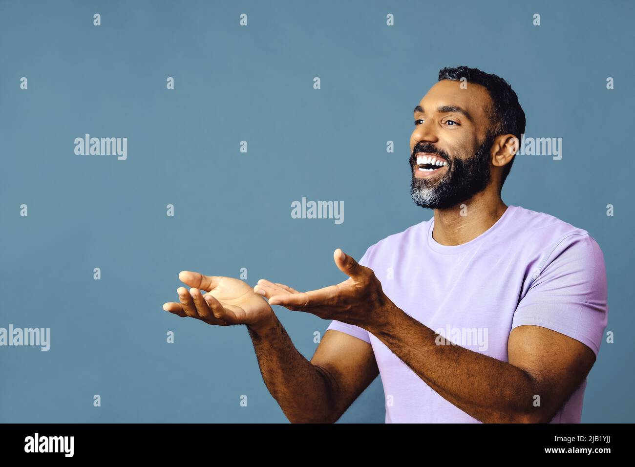 jeune homme noir avec barbe et moustache souriant et regardant loin de l'espace de copie main signal isolé de fond bleu studio Banque D'Images