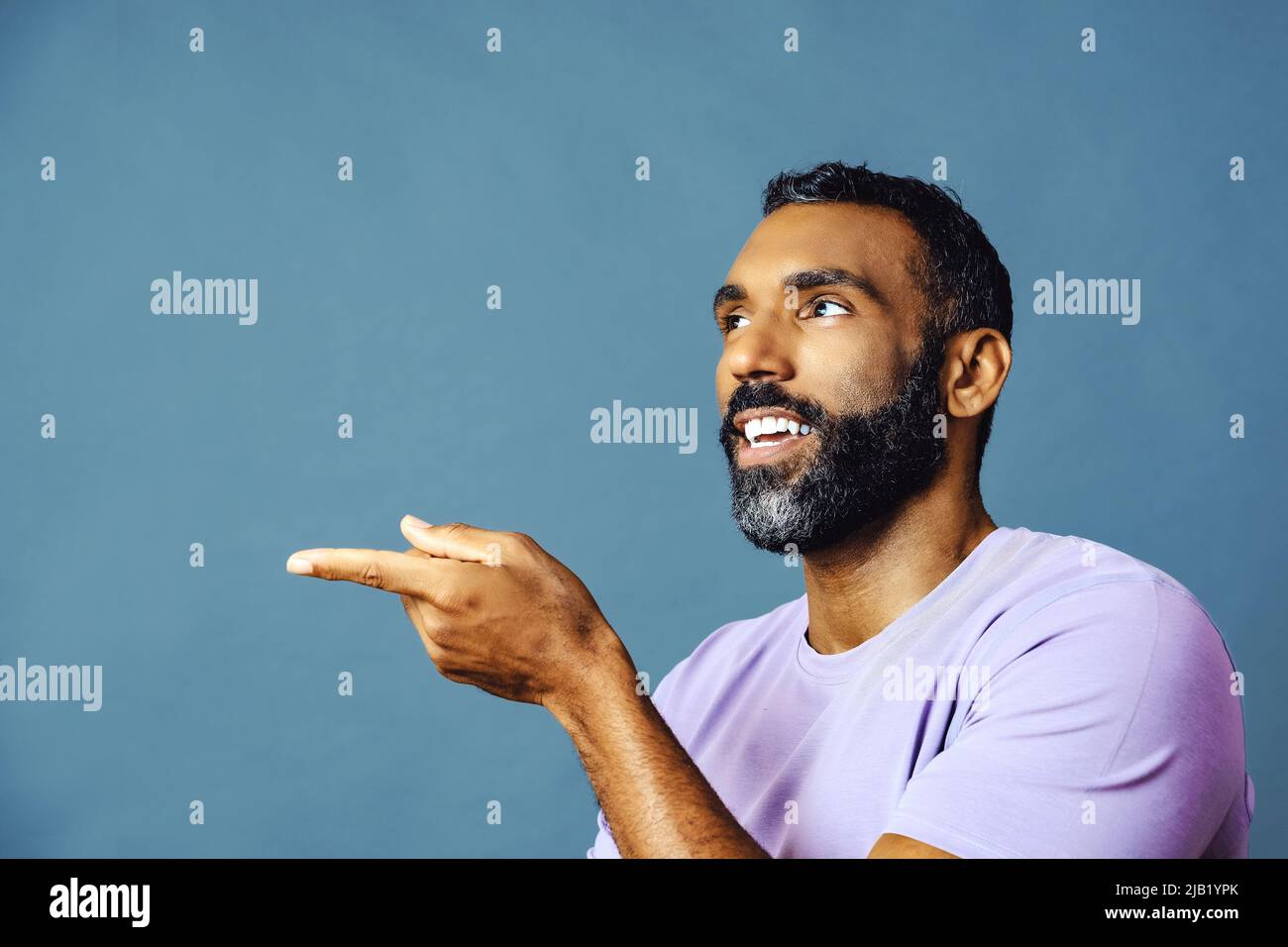 jeune homme noir avec barbe et moustache souriant et regardant loin de l'espace de copie main signal isolé de fond bleu studio Banque D'Images