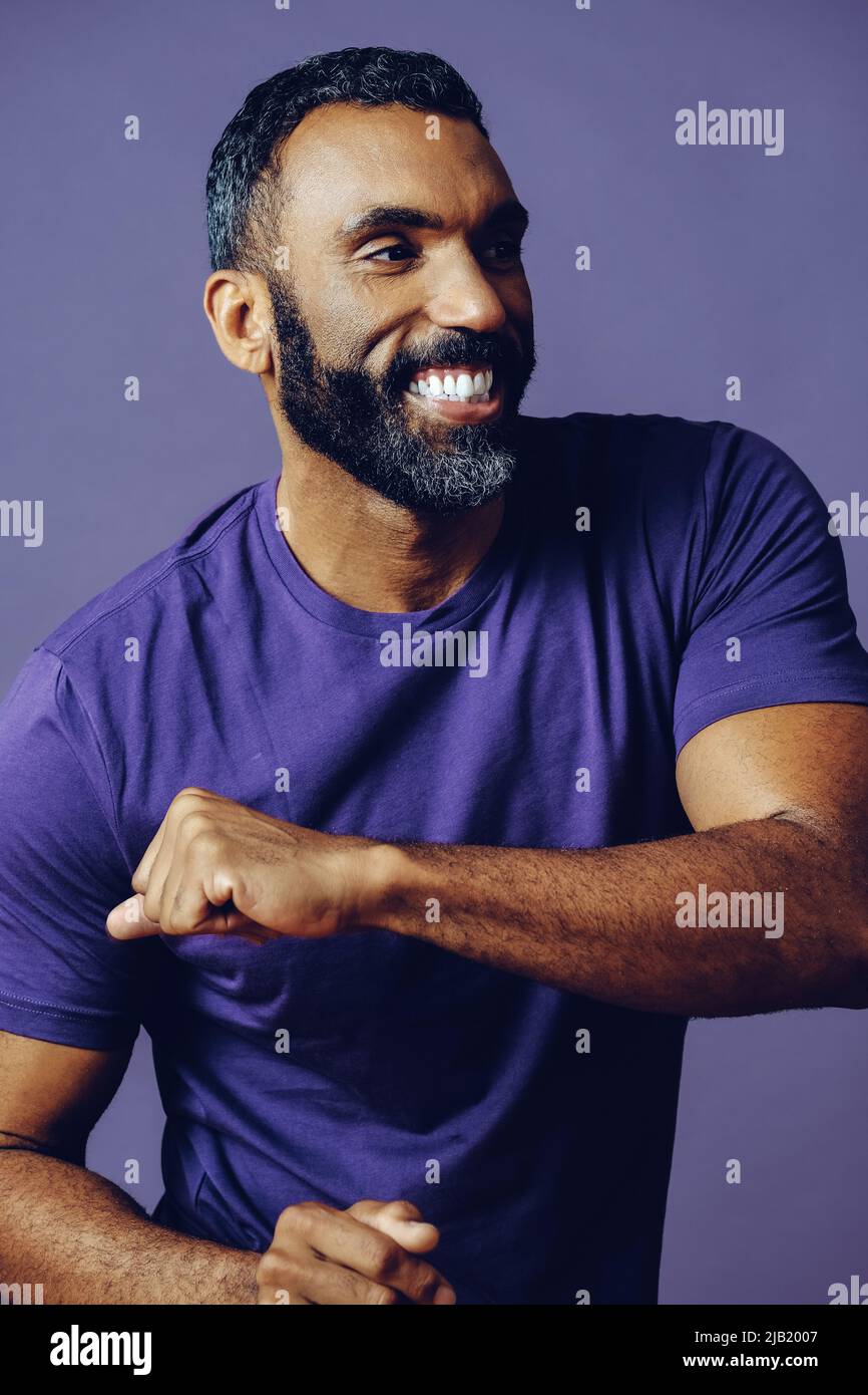 portrait d'un homme réussi dansant avec une barbe et un t-shirt violet célébrant avec bras vers le haut sur un fond violet studio Banque D'Images