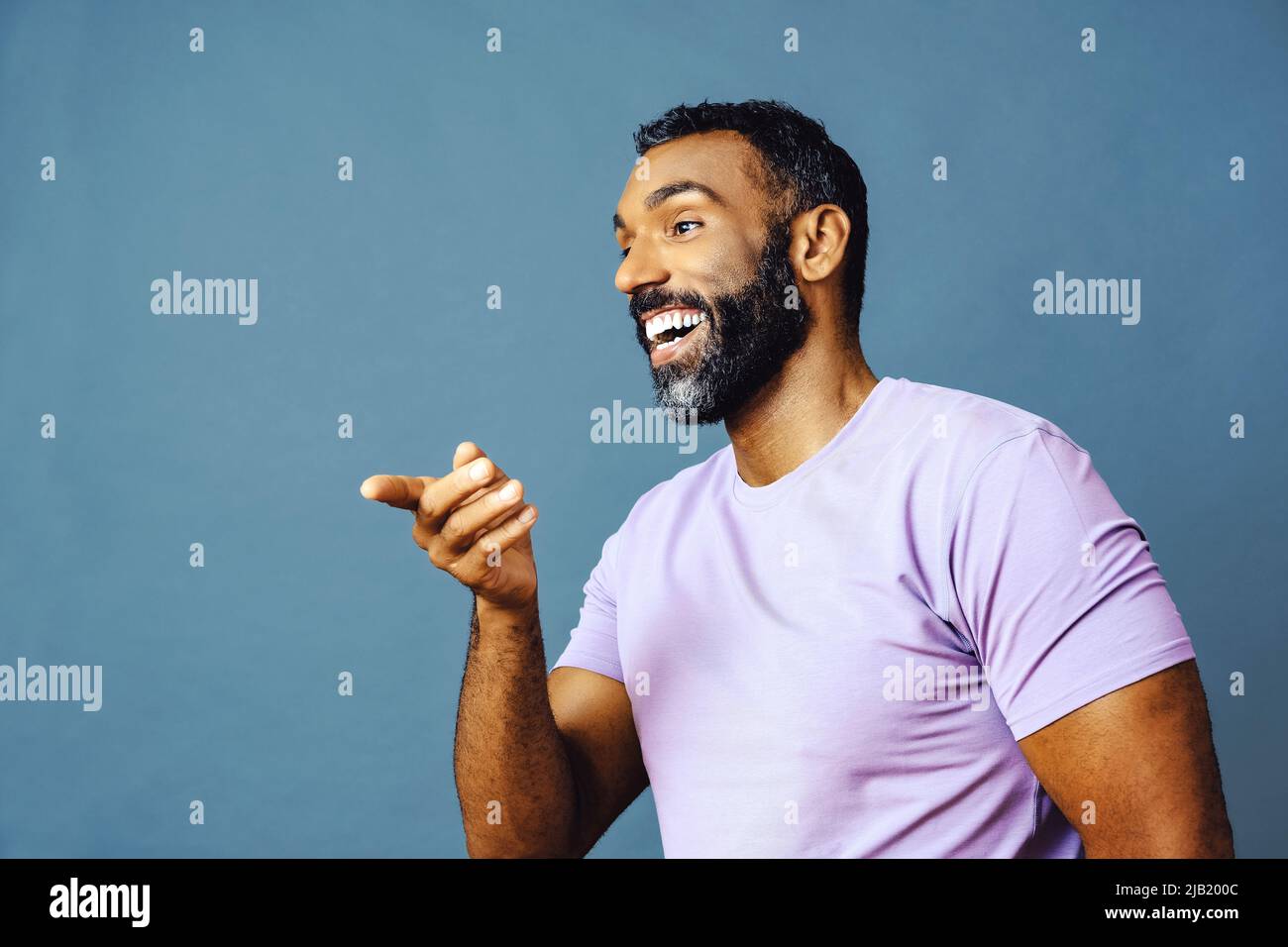 jeune homme noir avec barbe et moustache souriant et regardant loin de l'espace de copie main signal isolé de fond bleu studio Banque D'Images